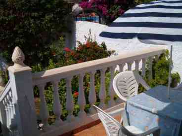 Sunny raised terrace furnished with table and chairs leads to a small mature garden. 