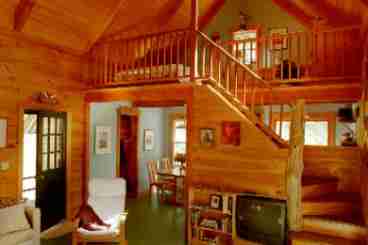 Viewed from across the living room is the dining room and carpeted loft bedroom. The compact but efficient kitchen is partly visible through the opening in the staircase. The interior is beautifully crafted with solid birch walls, black walnut and driftwood staircase and easy-care painted floors.